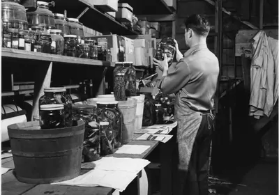 A black and white photo of a man standing at a lab bench, holding up a glass jar